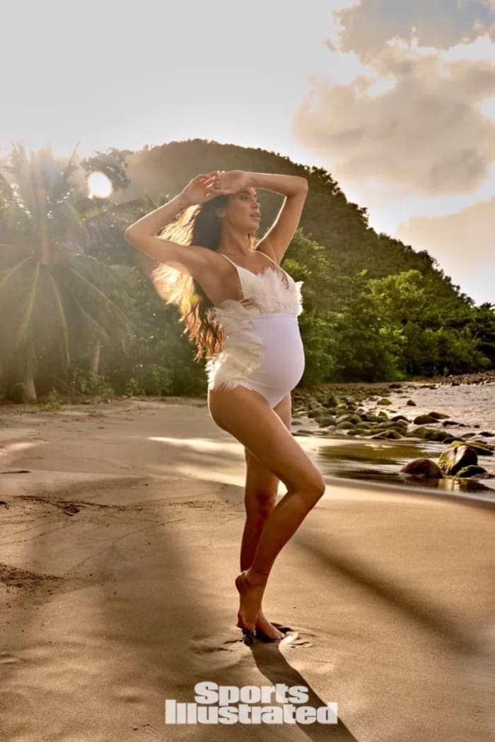 Nicole Williams English poses on the beach in front of the vibrant greenery in a white feathered one-piece.